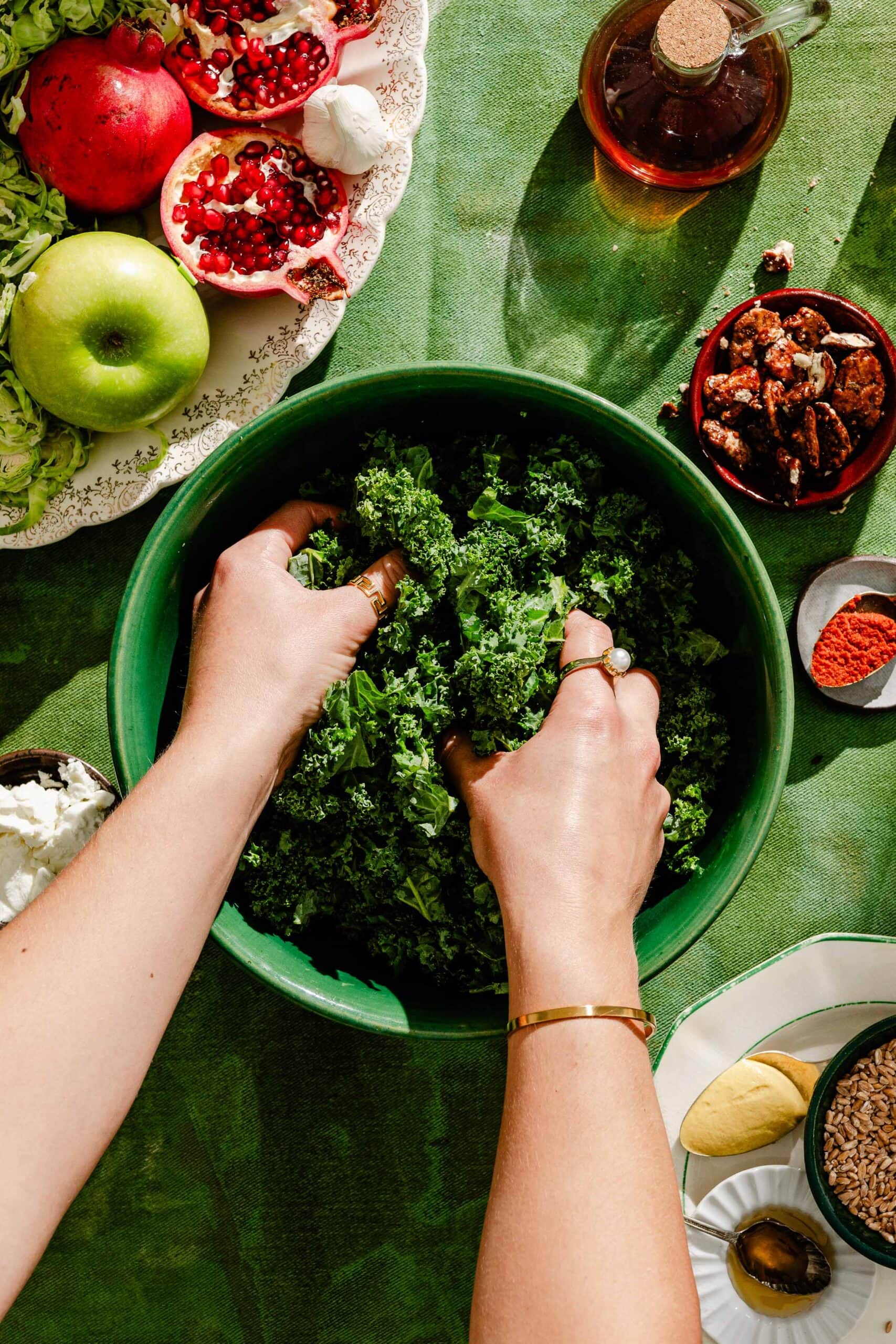 Hands tossing kale in a green bowl, surrounded by ingredients including pomegranates, an apple, pecans, cheese, and spices on a green tablecloth.