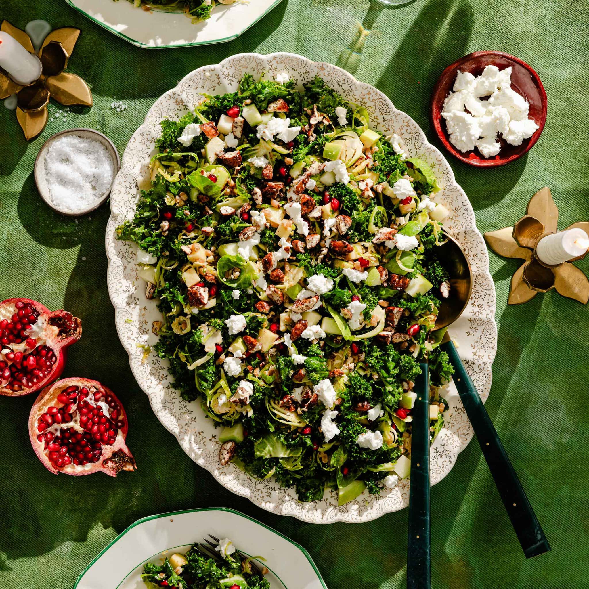A large platter of kale and Brussels sprout salad topped with pomegranate seeds, nuts, and goat cheese, served on a green tablecloth with sides of salt, extra cheese, and halved pomegranates.