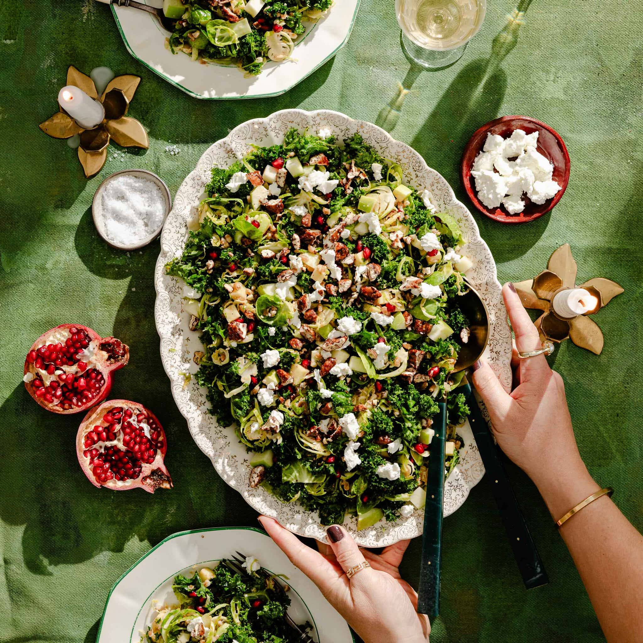 A large oval platter of kale and Brussels sprout salad topped with pomegranate seeds, nuts, and crumbled cheese, being served at a table with halved pomegranates and small bowls of salt and cheese.