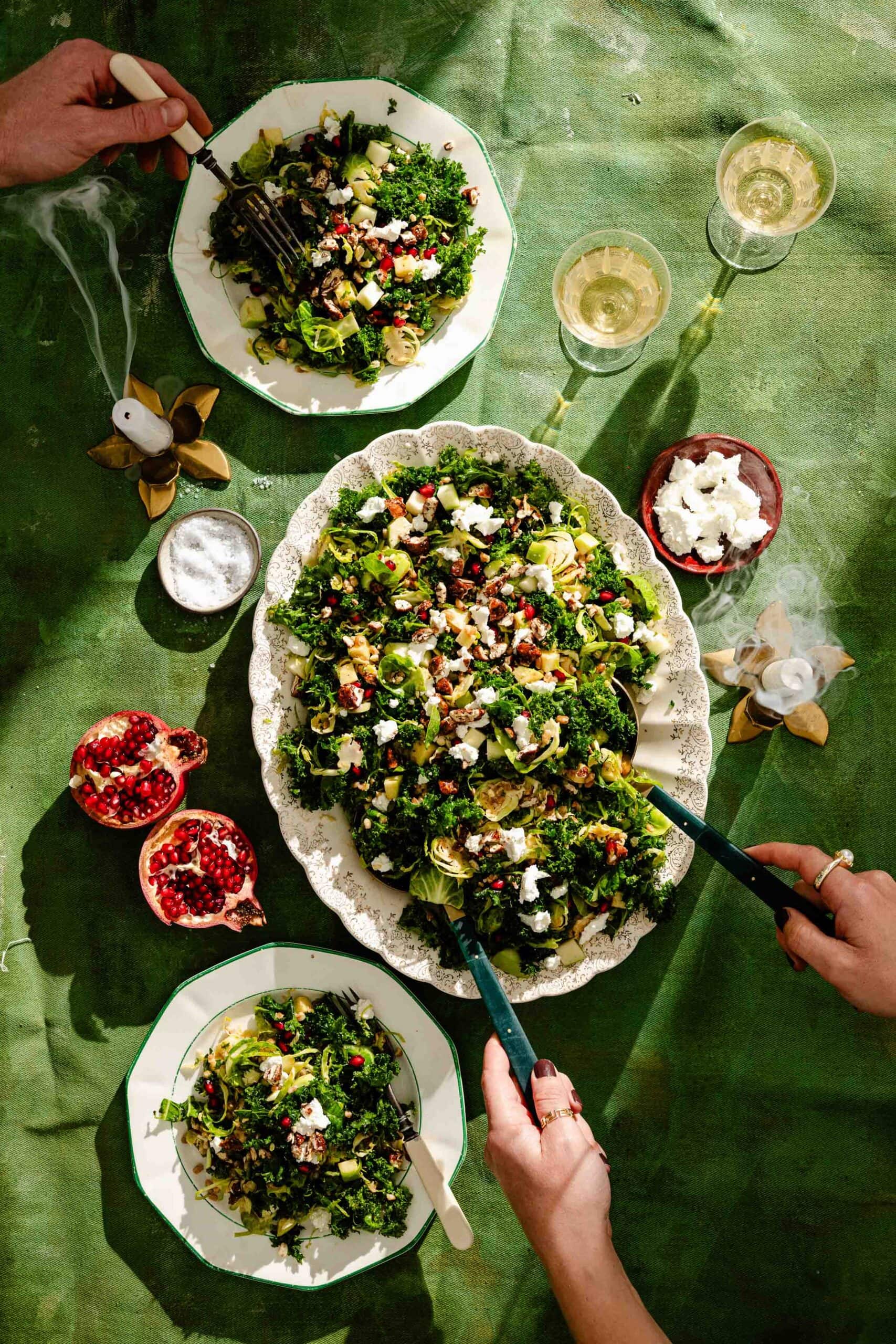 A large platter of kale and Brussels sprout salad topped with pomegranate seeds and crumbled cheese, surrounded by individual servings, two glasses of white wine, and halved pomegranates on a green tablecloth.