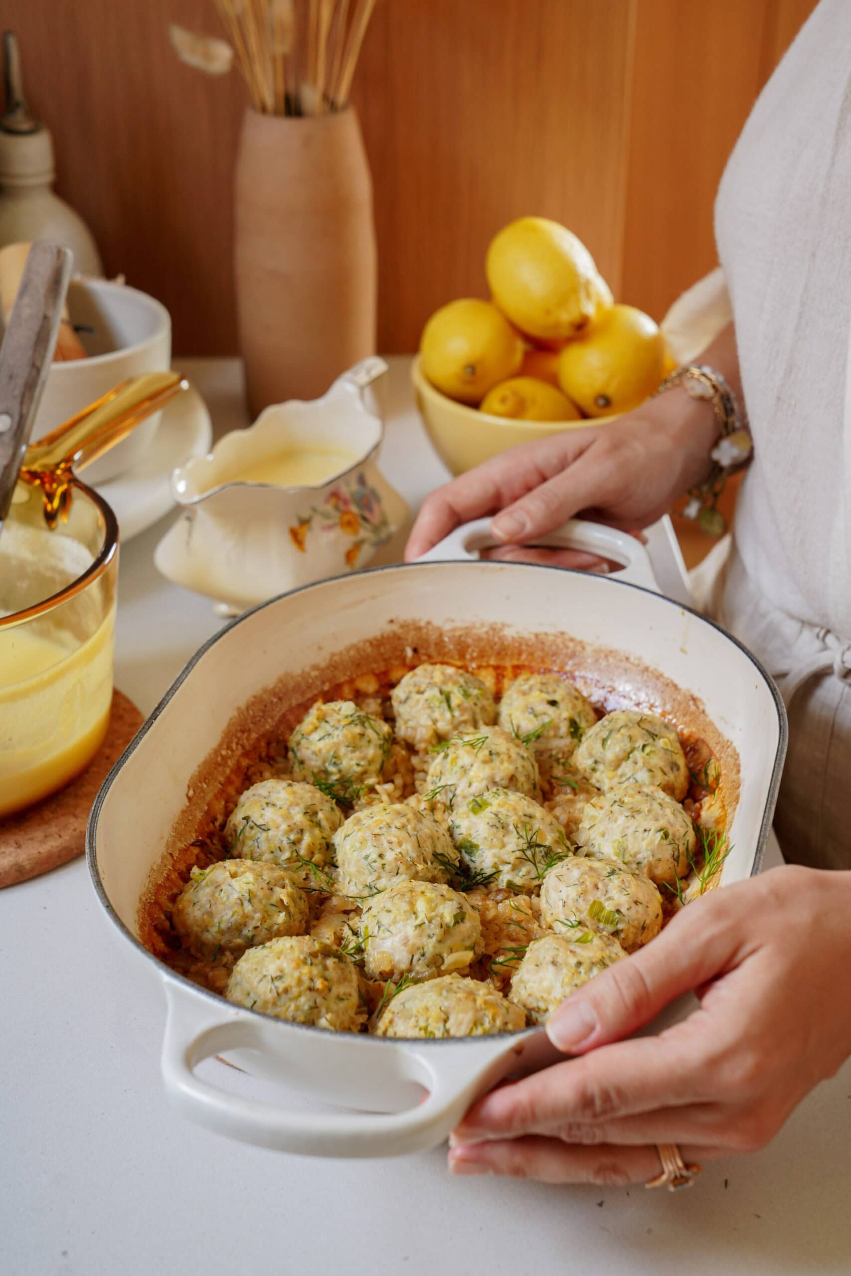 A person holds a shallow baking dish filled with herb-topped meatballs, with lemons and a gravy boat in the background.