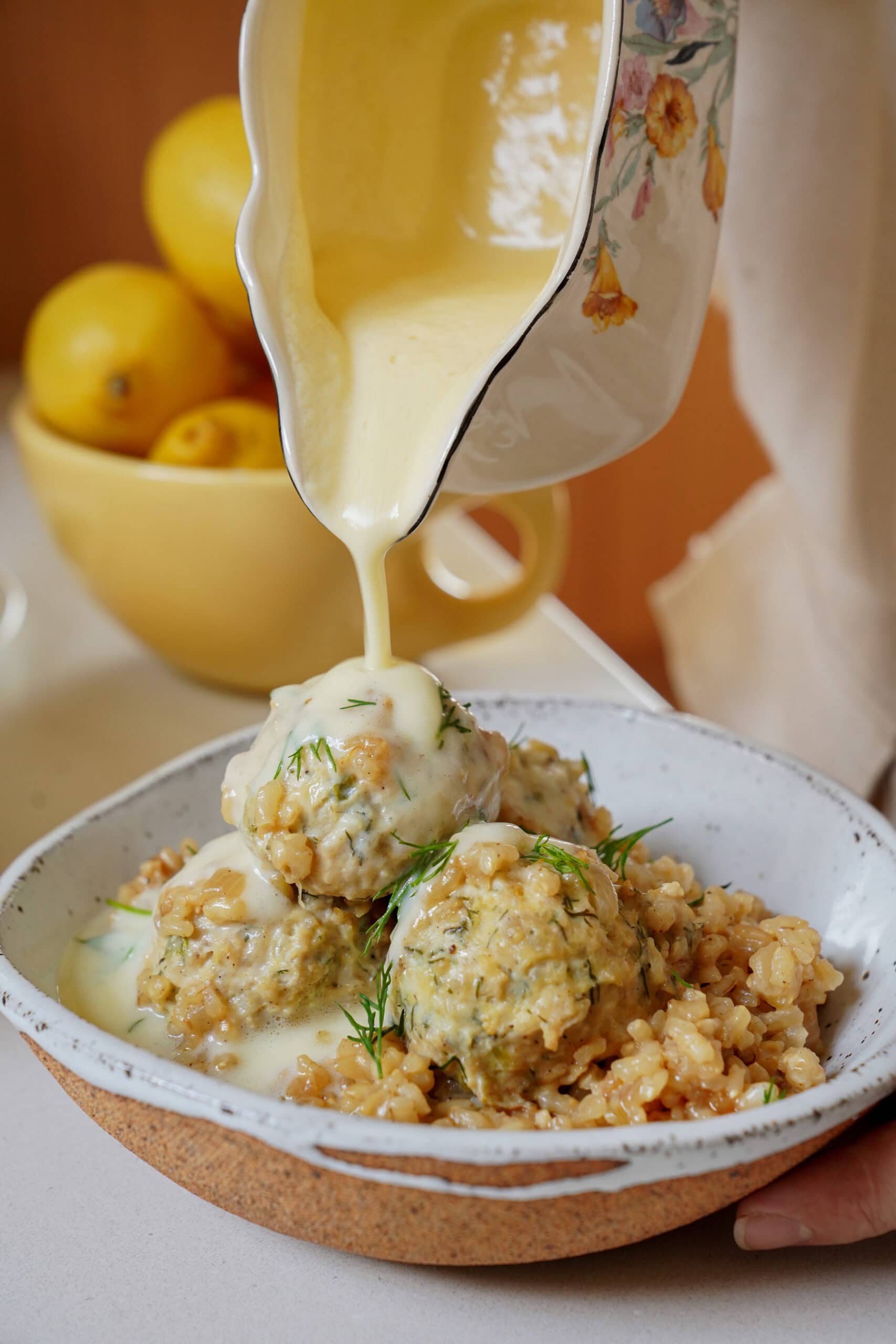 Creamy lemon sauce being poured from a floral gravy boat over a bowl of Greek-style meatballs and rice garnished with dill.