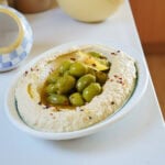 A bowl of hummus topped with green olives and red pepper flakes.