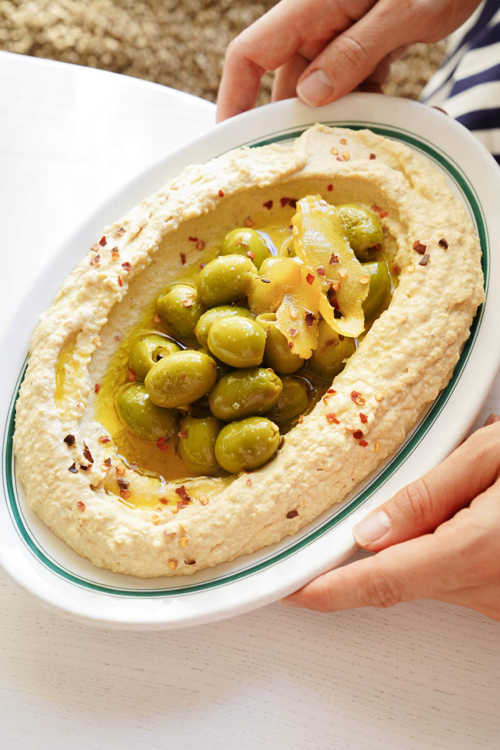 Oval platter of hummus topped with green olives, lemon zest, and red pepper flakes, held by two hands.