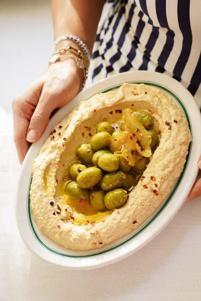 Hands holding an oval platter of hummus topped with green olives, olive oil, and red pepper flakes.