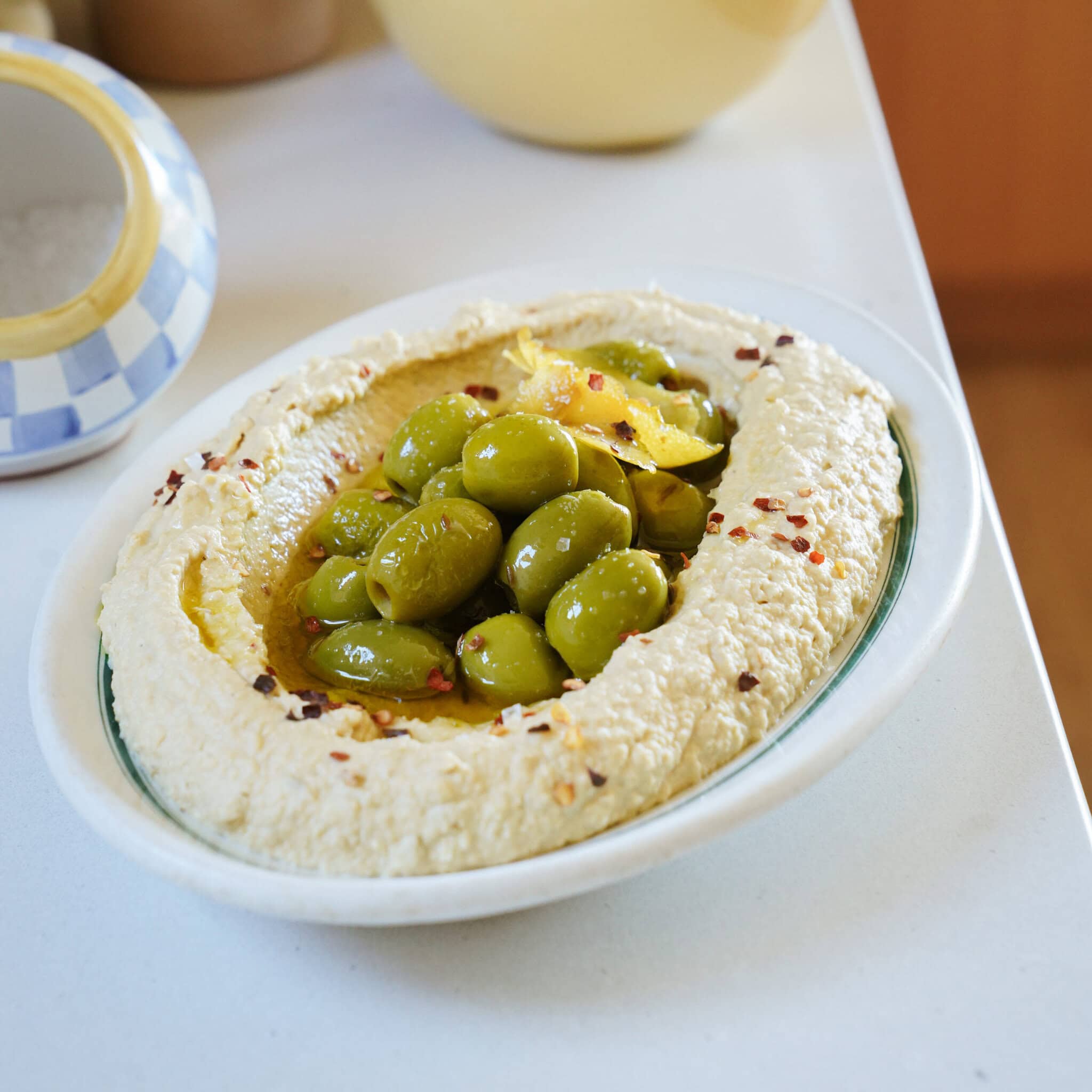 A bowl of hummus topped with green olives and red pepper flakes.