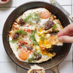 A hand dips a piece of toast into a cast-iron skillet containing fried eggs topped with dill, spices, and dates.