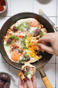 A cast-iron skillet containing fried eggs topped with dates, spices, and dill, with a hand dipping a piece of bread into the runny yolk.