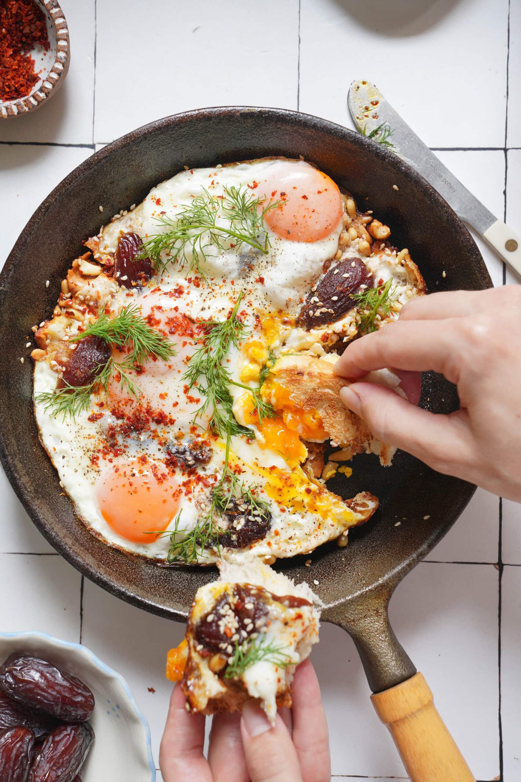A cast-iron skillet containing fried eggs topped with dates, spices, and dill, with a hand dipping a piece of bread into the runny yolk.