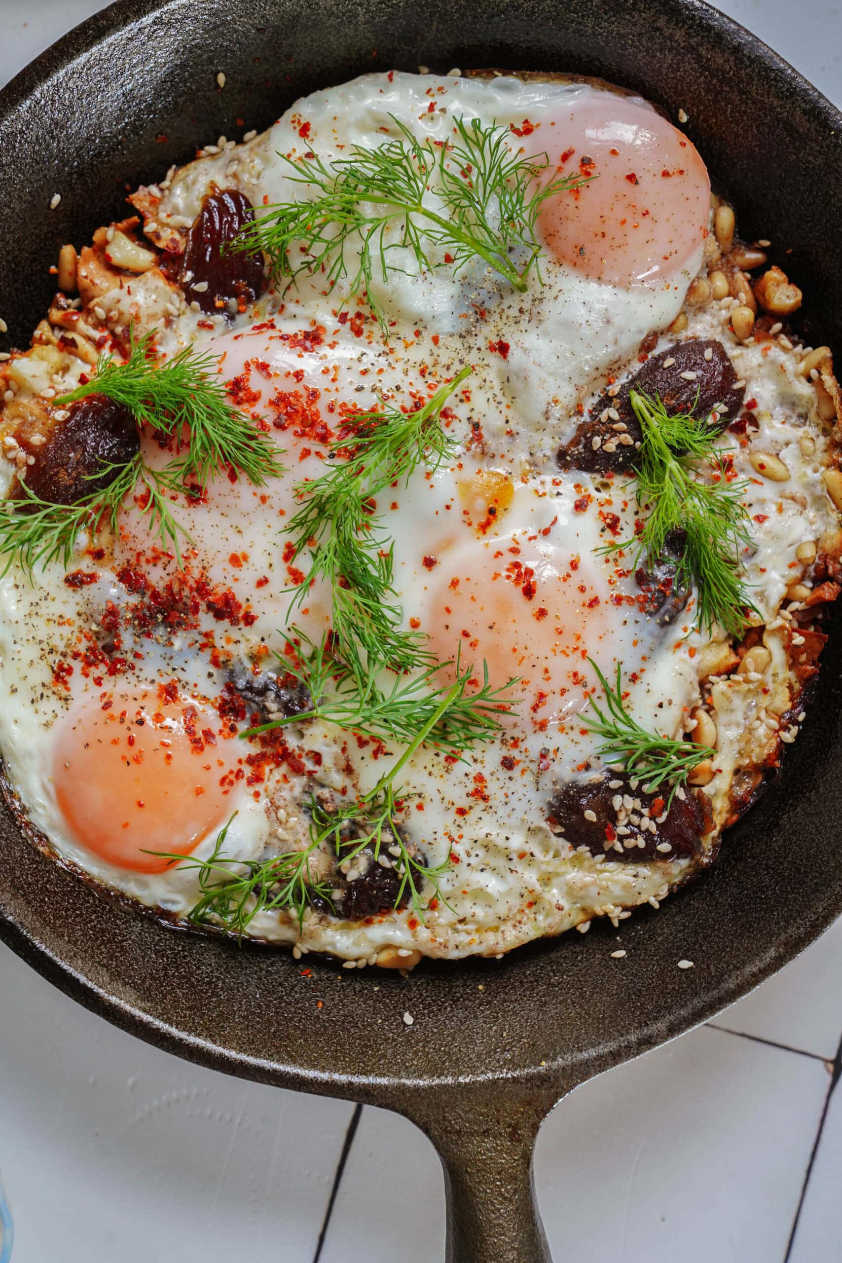 Three fried eggs in a cast-iron skillet, garnished with fresh dill, red pepper flakes, sesame seeds, and dark fruit pieces.