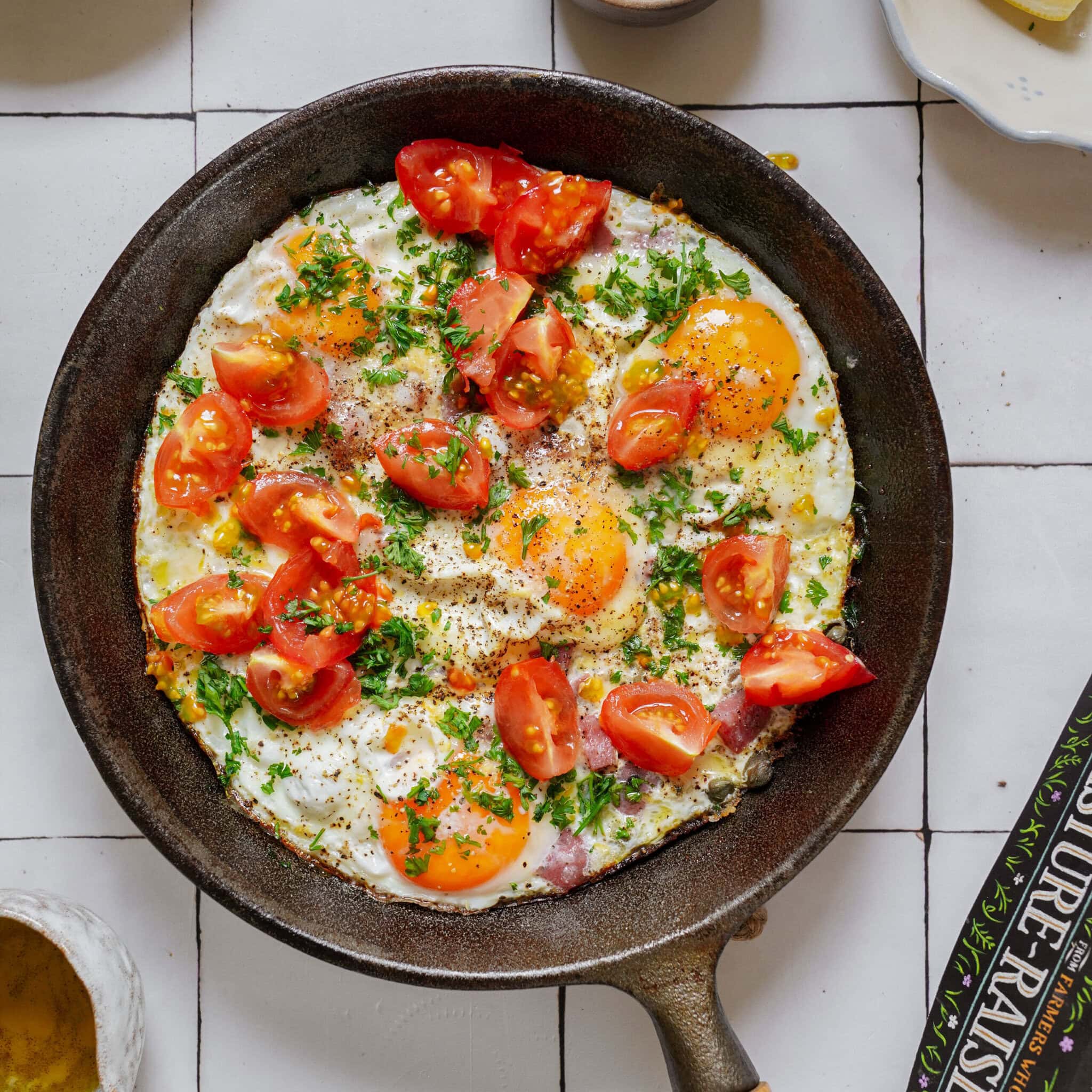 Cast iron skillet containing fried eggs topped with halved cherry tomatoes and chopped herbs.