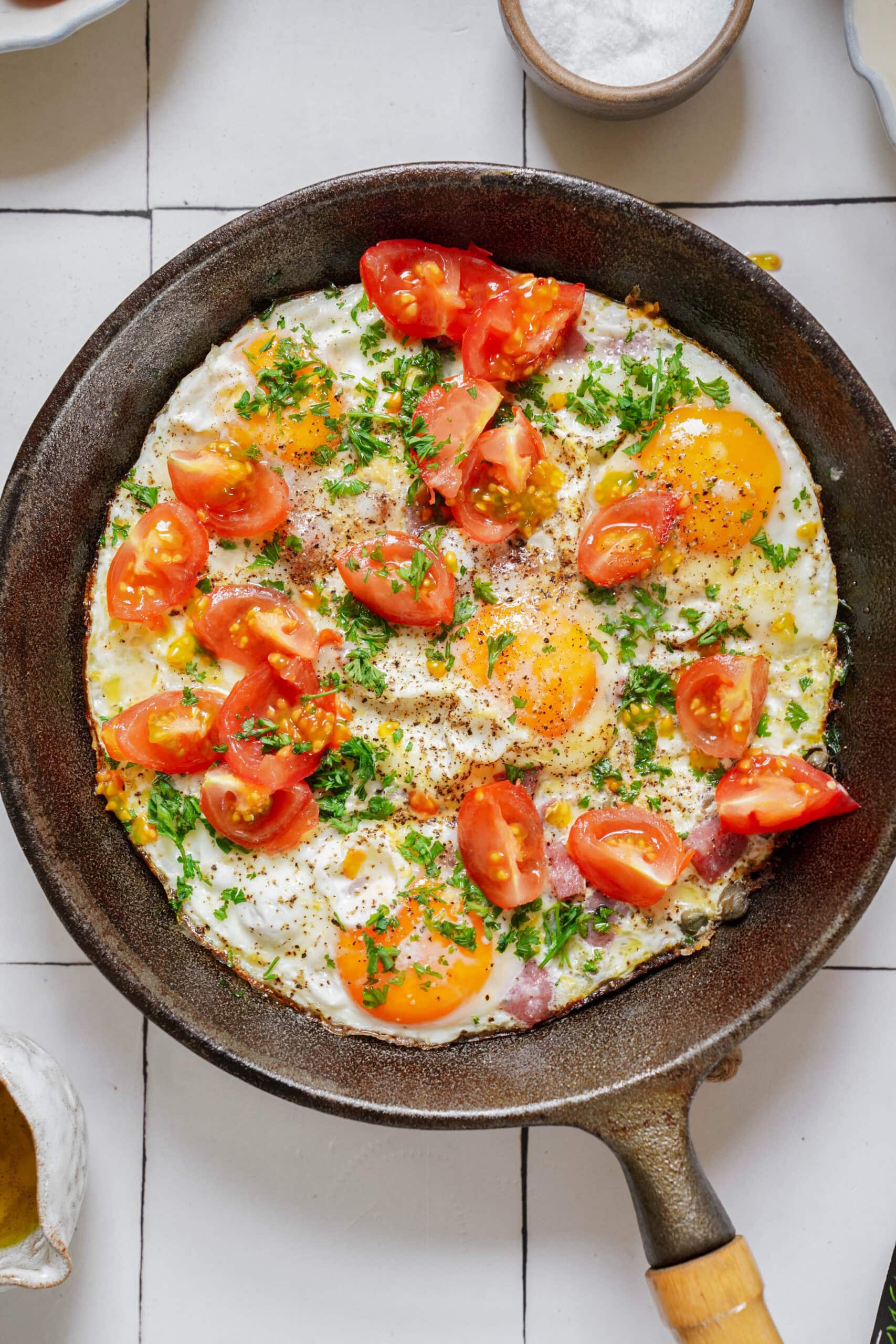Skillet of fried eggs topped with halved cherry tomatoes and fresh herbs.