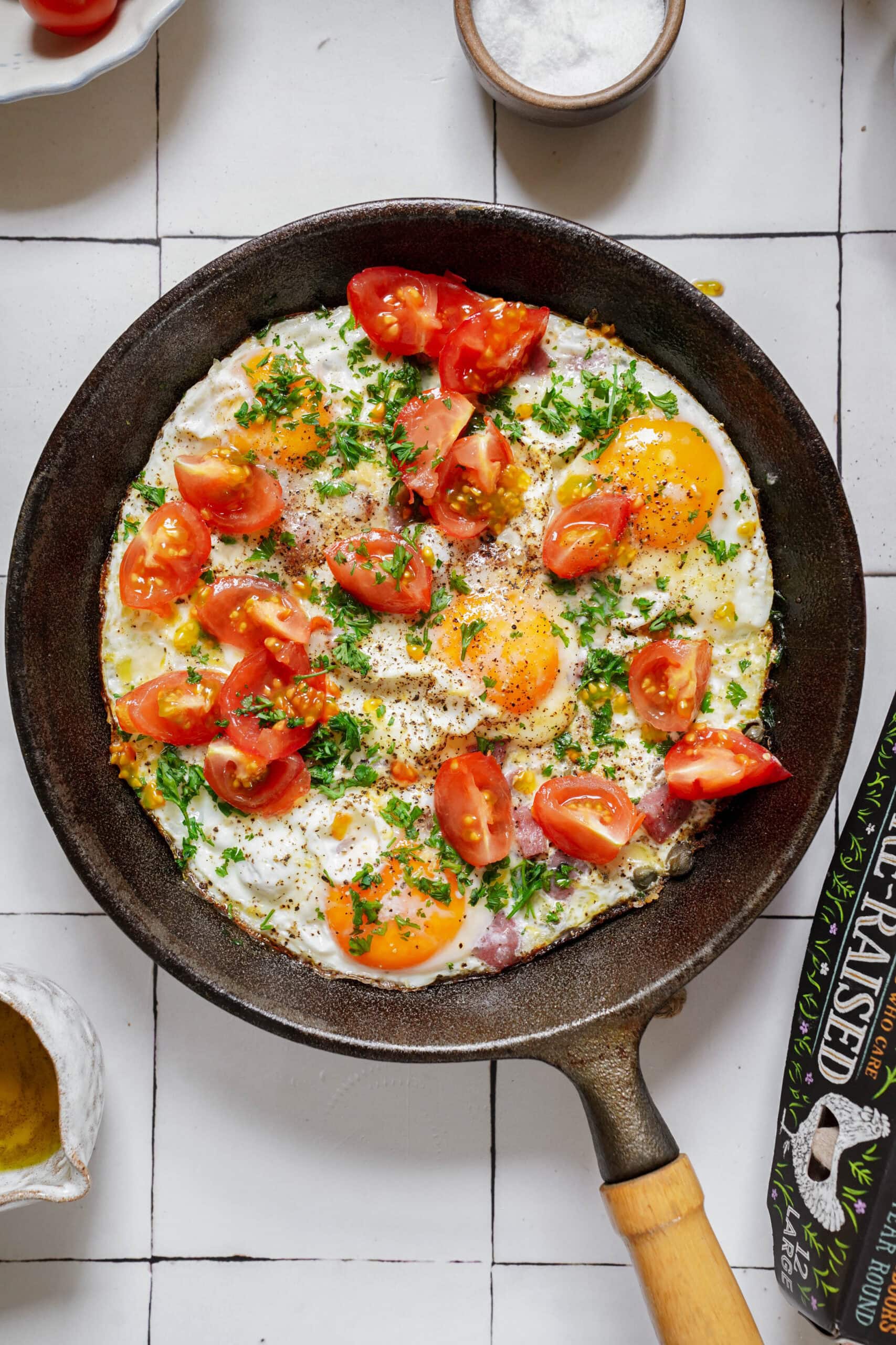 Fried eggs in a cast iron skillet topped with cherry tomatoes, fresh herbs, and black pepper.