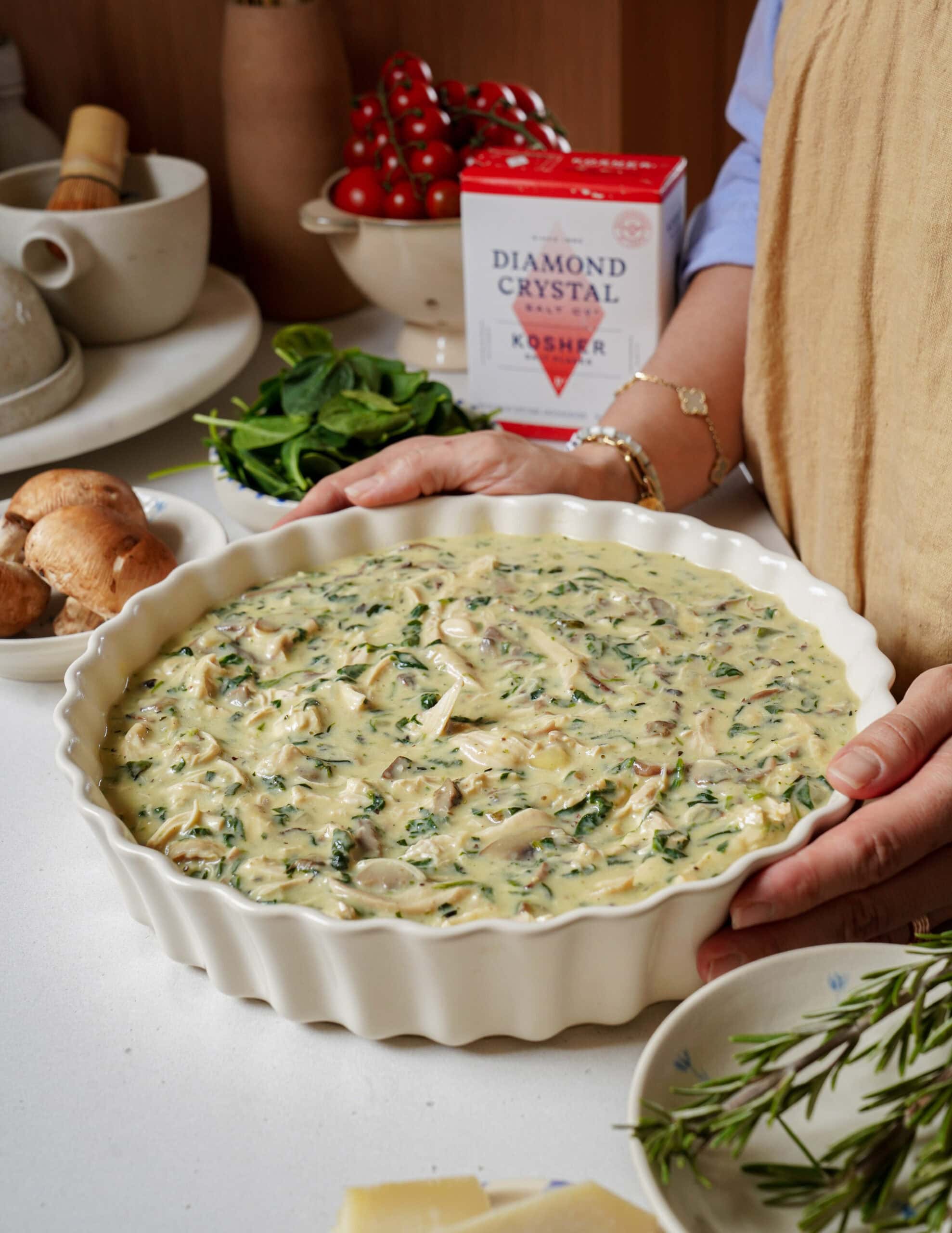 A person holding a large ceramic baking dish filled with an unbaked creamy chicken, spinach, and mushroom mixture.