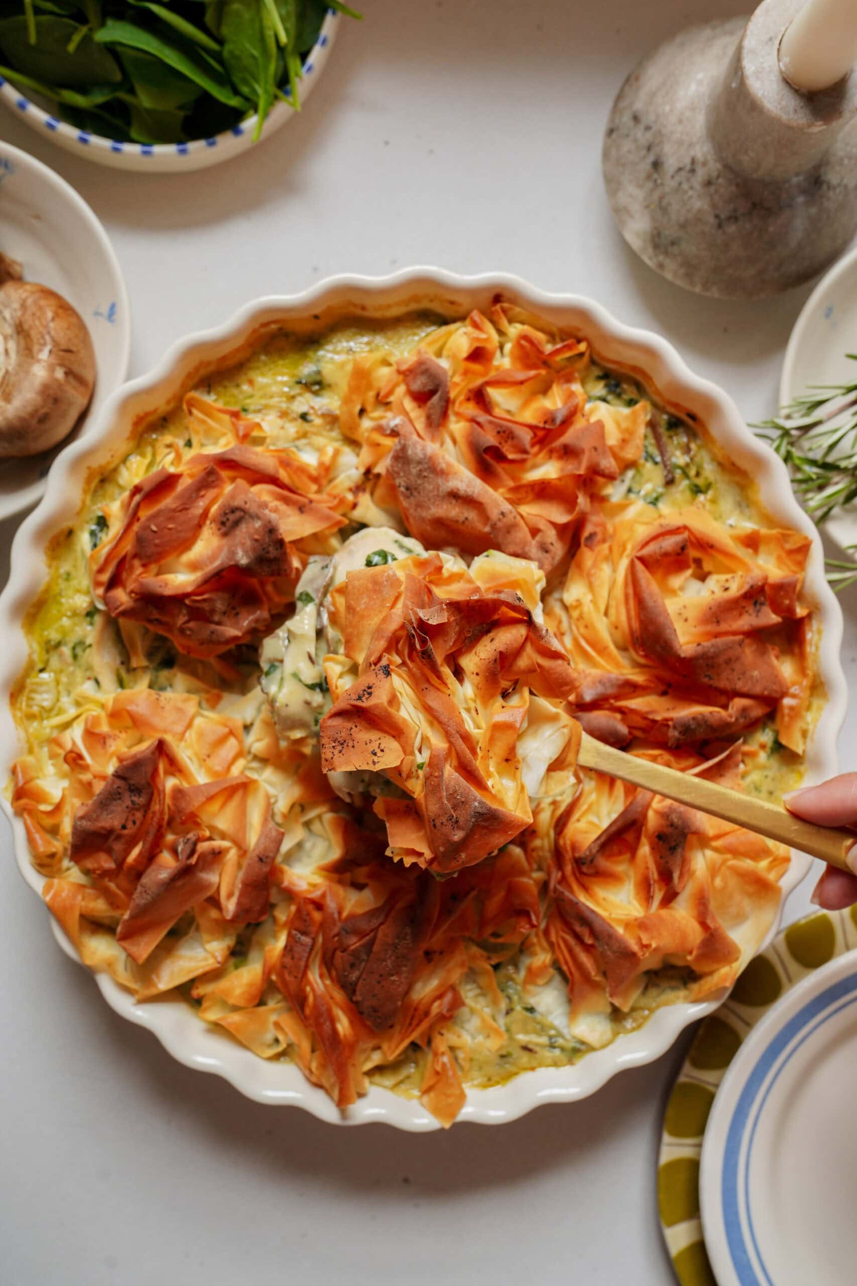 A hand uses a wooden spoon to lift a portion of spinach and phyllo pie from a ceramic baking dish.