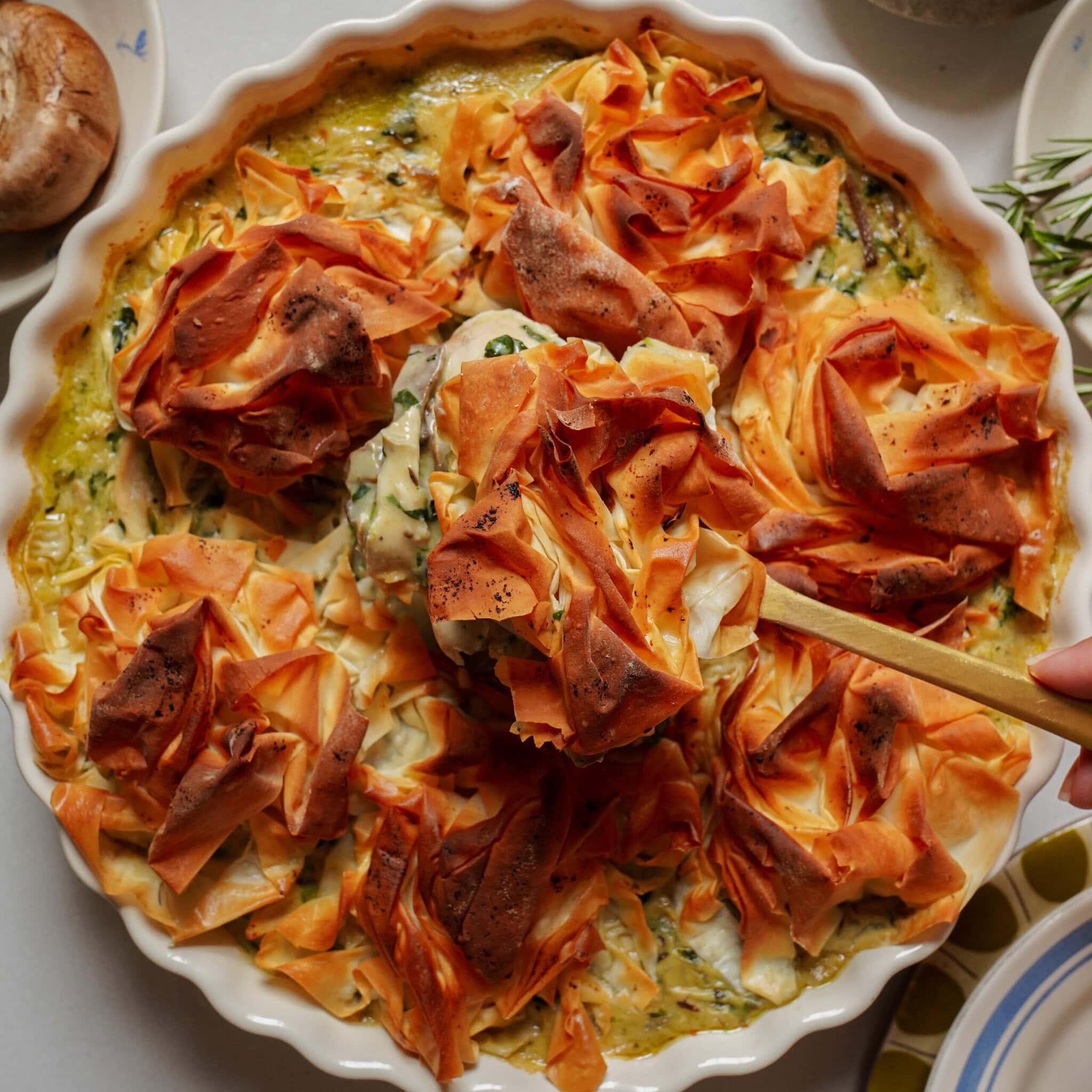 A hand uses a wooden spoon to lift a serving of savory pie topped with golden, ruffled phyllo pastry from a round baking dish.