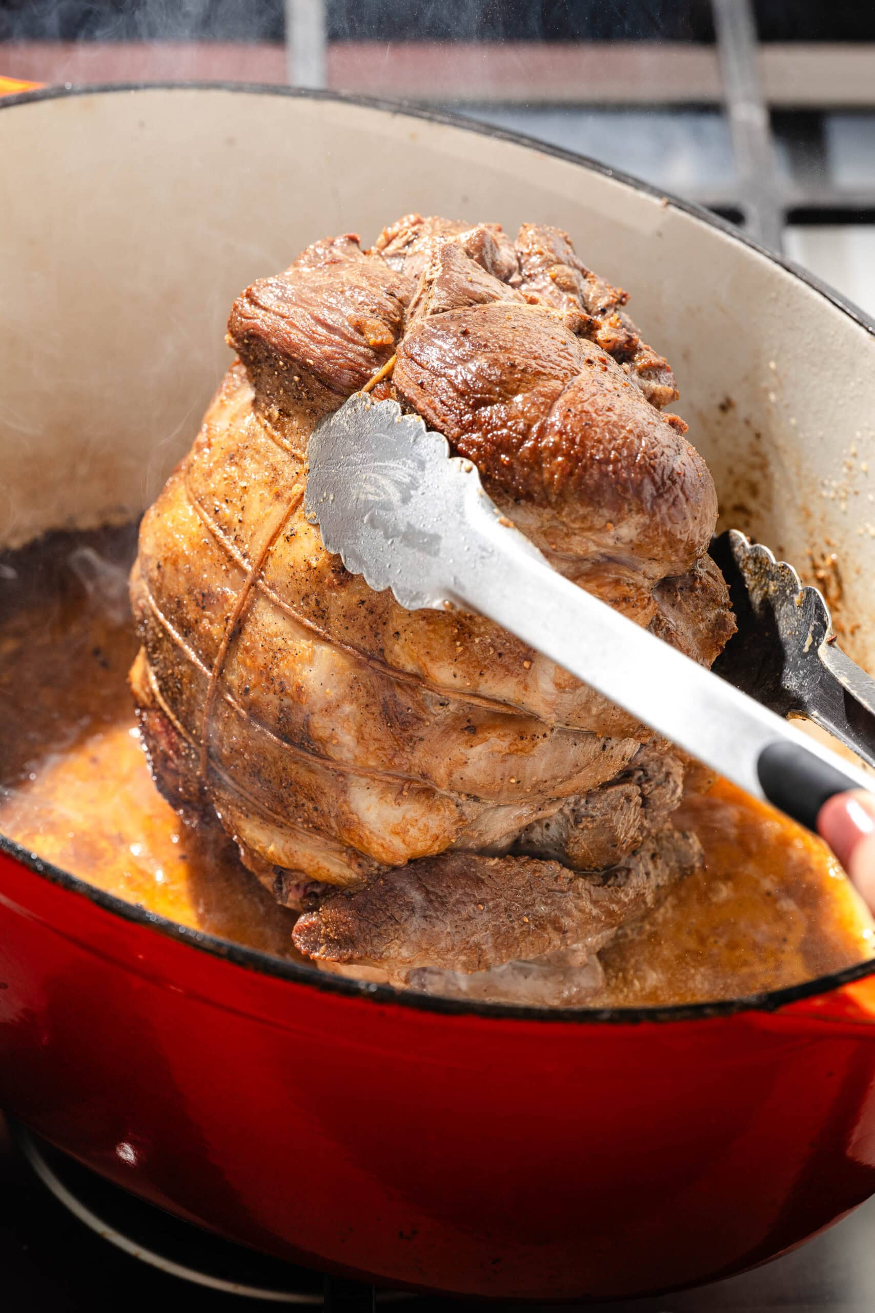 Metal tongs lifting a seared, kitchen-twined roast from a red enameled cast iron pot.