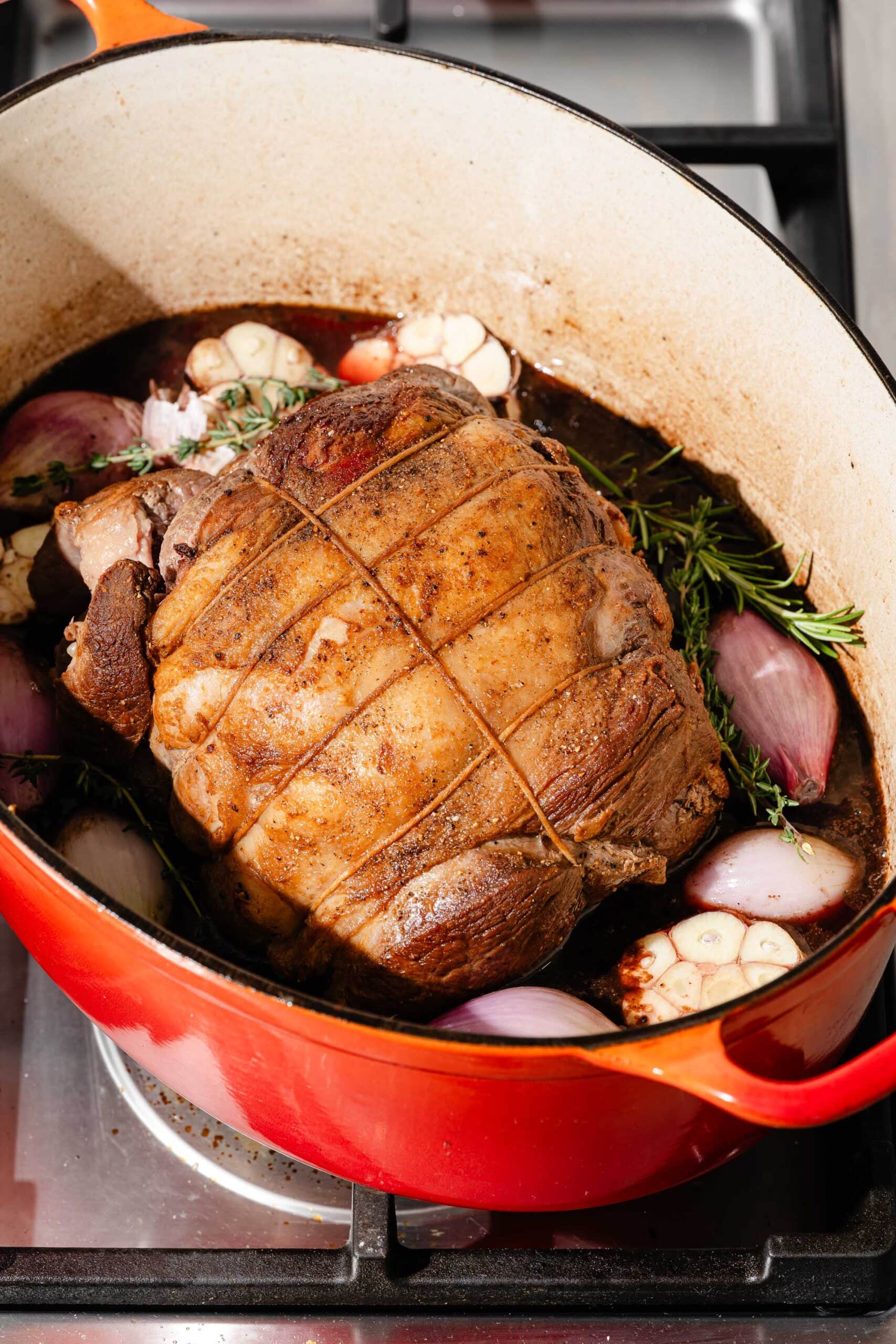 A tied roast in a red Dutch oven with garlic cloves, red onion, and fresh herbs in a dark cooking liquid.