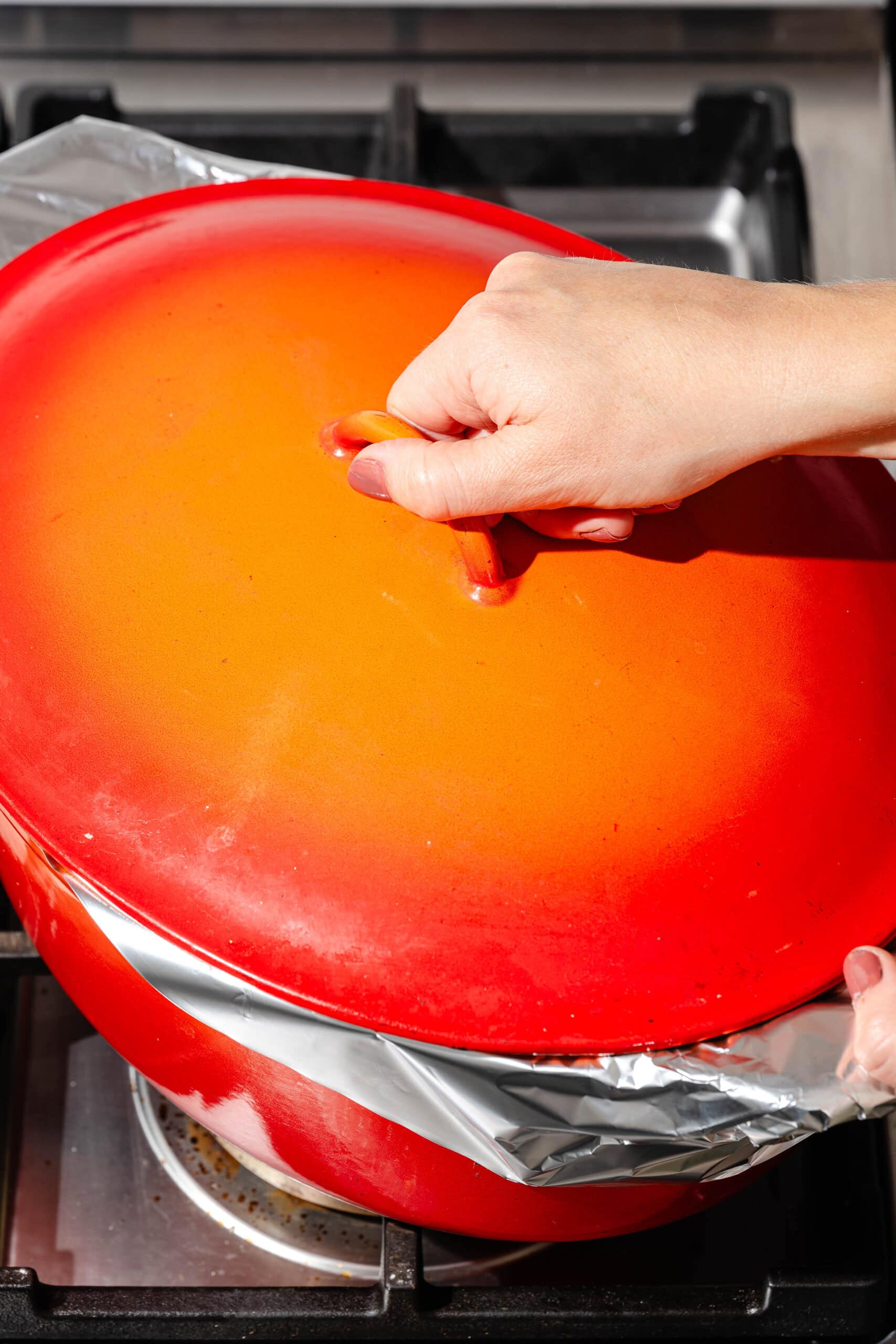 A hand lifts the bright red lid of a Dutch oven sealed with a layer of aluminum foil on a stovetop.