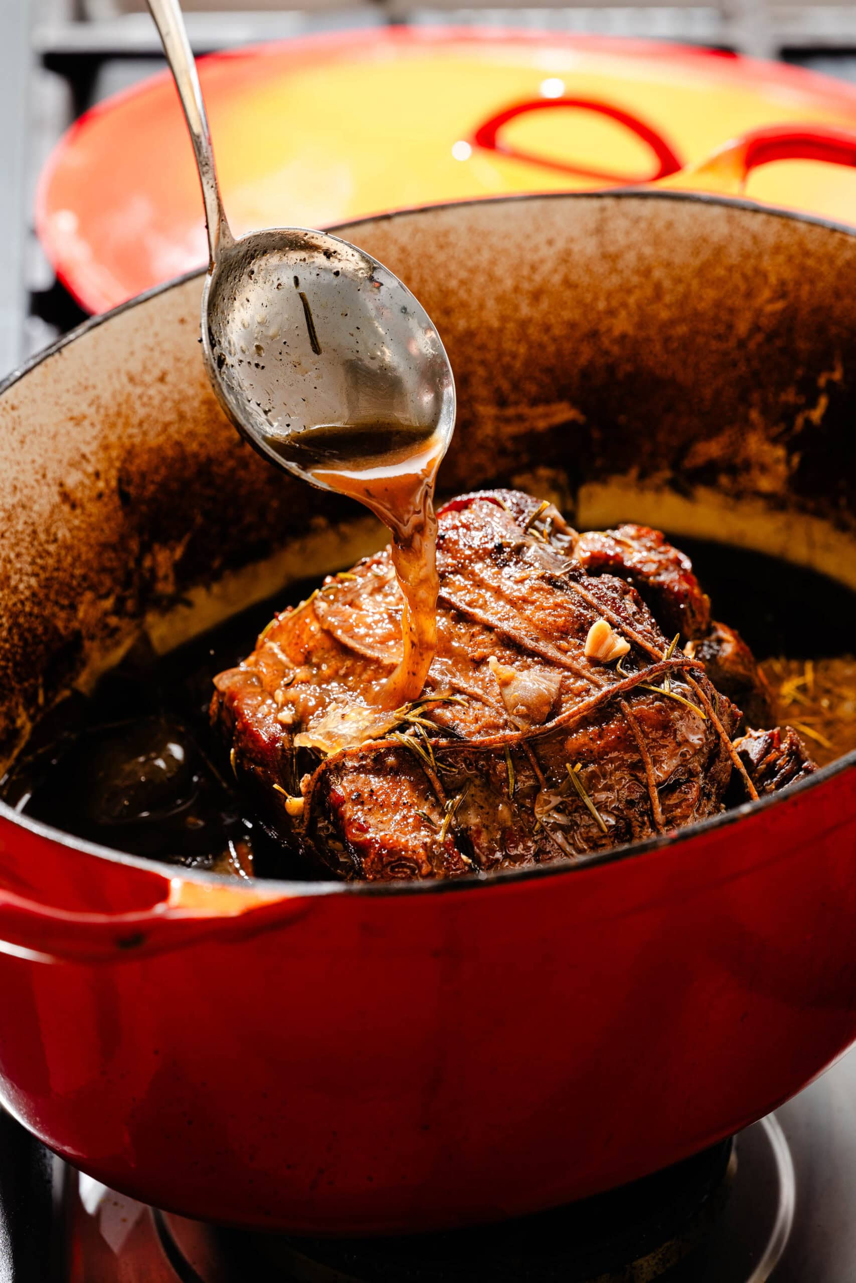 A ladle pours juices over a roasted meat dish inside a red enameled cast iron pot.