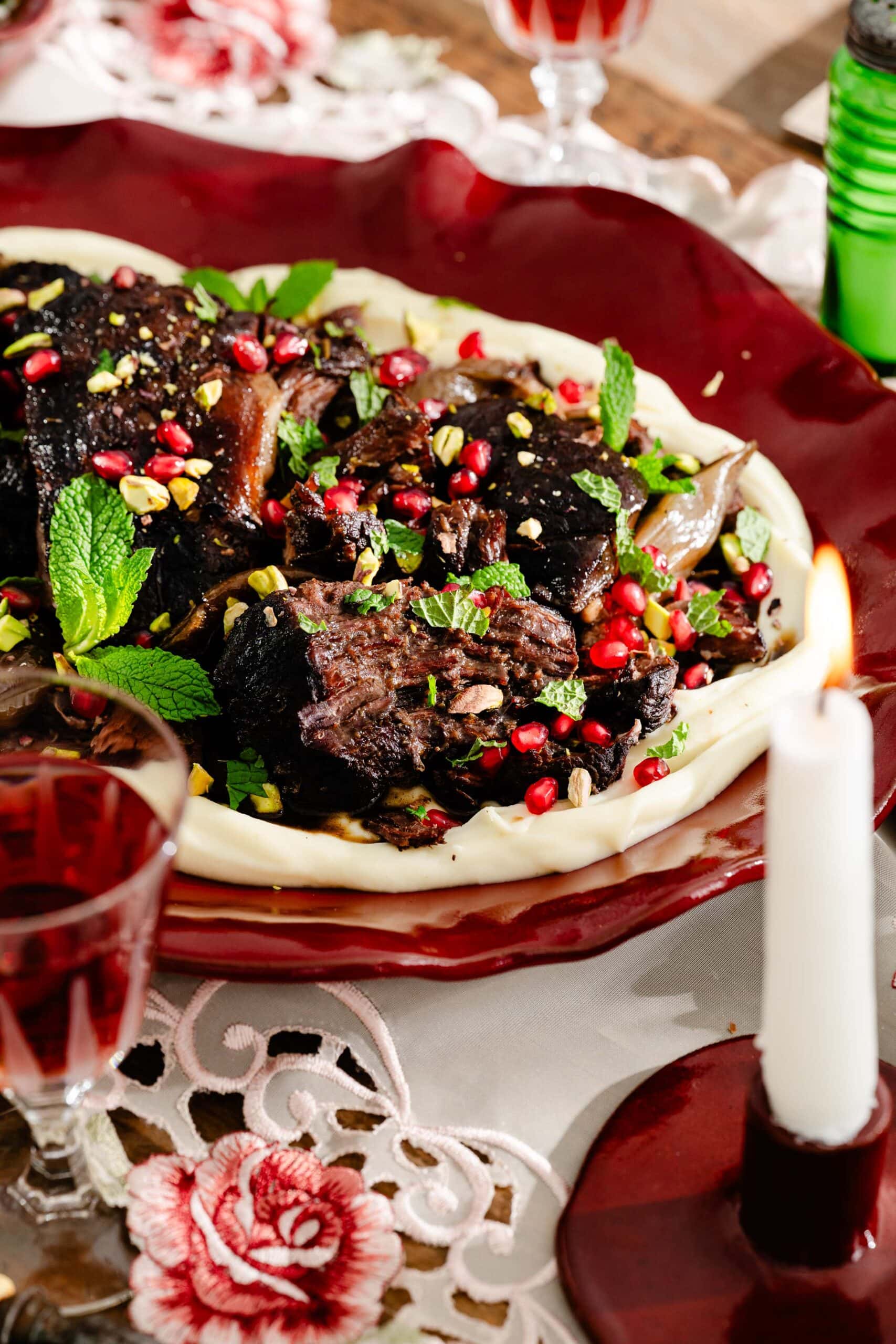 A platter of braised meat served over a bed of white puree, garnished with pomegranate seeds, chopped pistachios, and fresh mint.