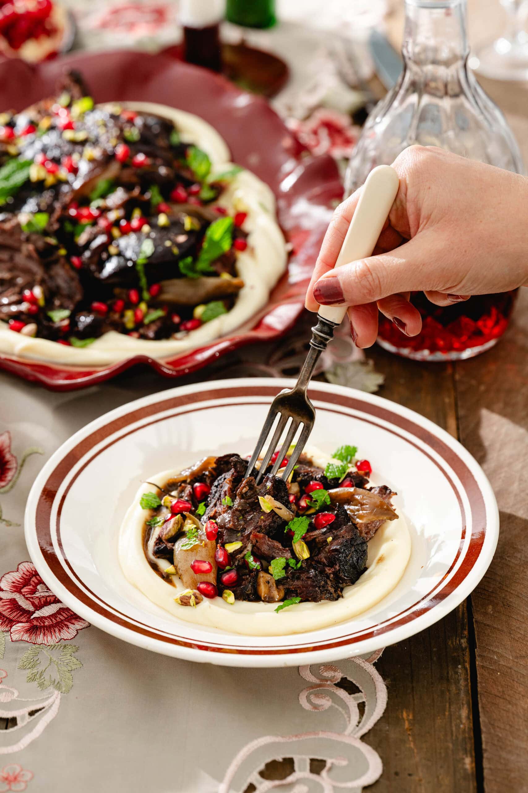 A hand uses a fork to lift a piece of braised meat from a plate of pureed potatoes topped with pomegranate seeds and herbs.