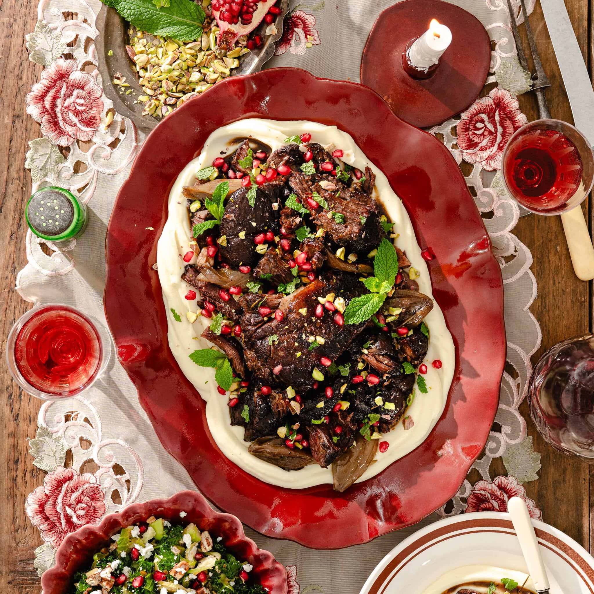 Braised beef short ribs served over a bed of creamy purée, topped with pomegranate seeds, chopped pistachios, and fresh mint on a red platter.