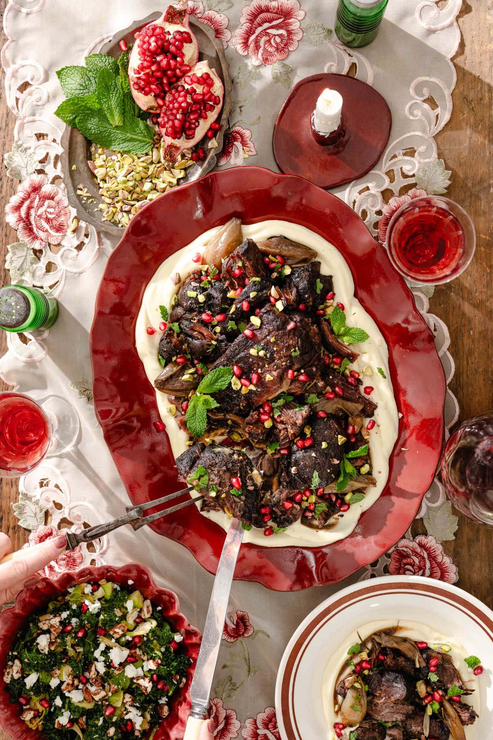A large platter of braised meat over pureed vegetables, garnished with pomegranate seeds and mint, set on a table with side dishes and wine.